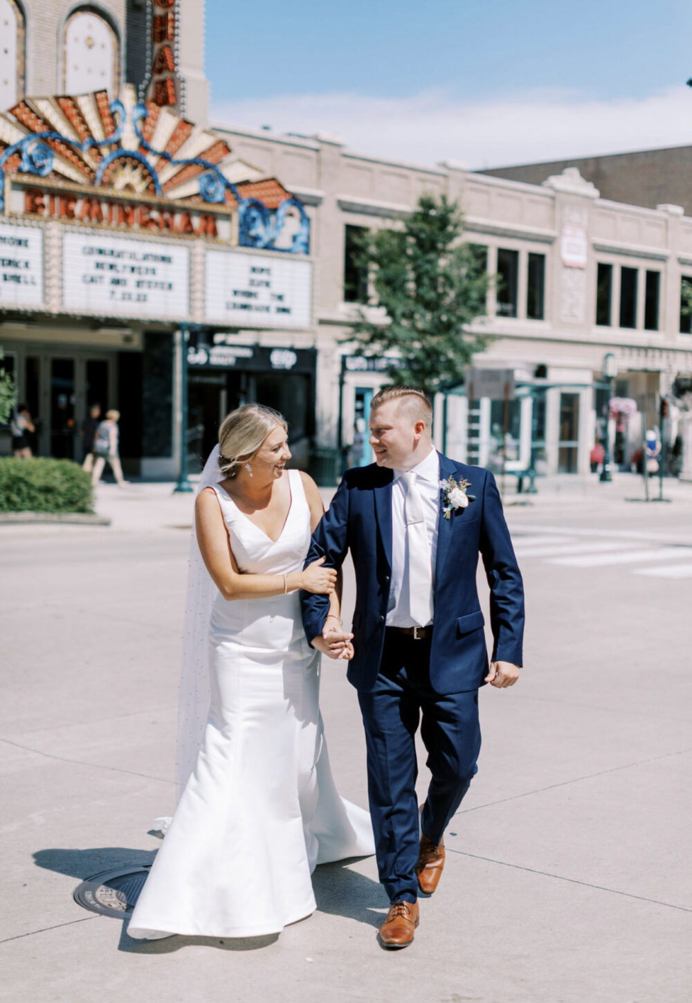 Bride and groom walking hand in hand across a downtown street in front of a historic theater marquee on their wedding day.