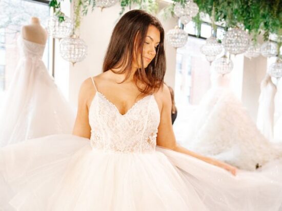 Bride in a sparkling tulle ball gown with a lace bodice stands in a bridal boutique beneath hanging crystal chandeliers and greenery.
