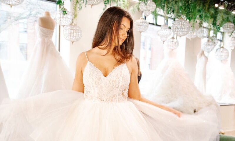Bride in a sparkling tulle ball gown with a lace bodice stands in a bridal boutique beneath hanging crystal chandeliers and greenery.