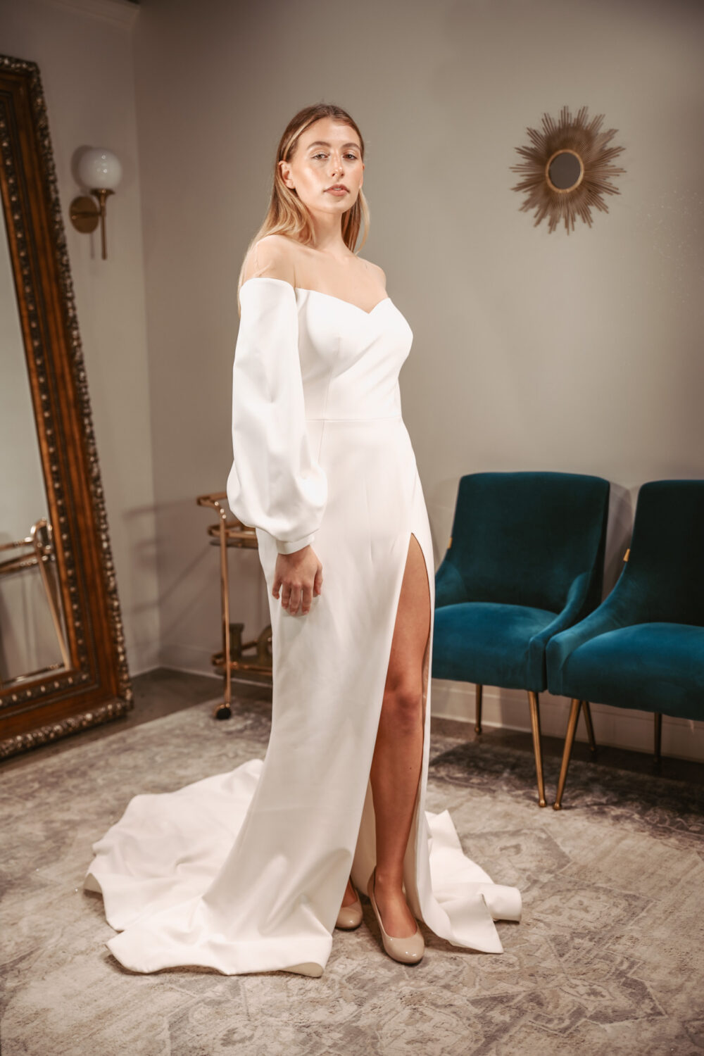 Woman wearing an off-the-shoulder wedding gown with long sleeves and a thigh-high slit stands beside teal velvet chairs in a bridal boutique.