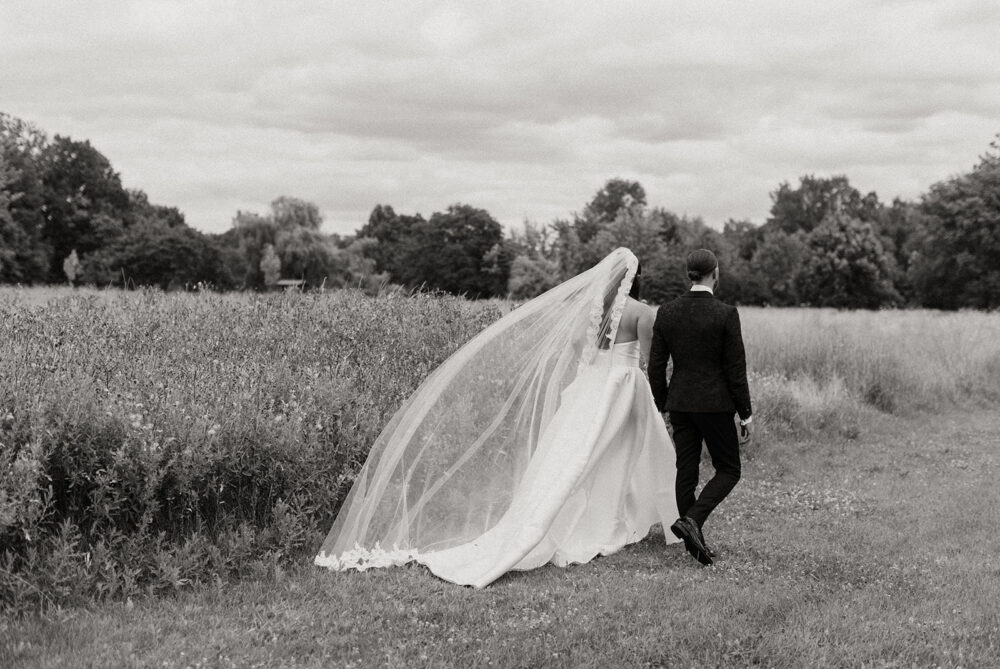 Bride and groom walking through a grassy field, with the bride’s long veil flowing behind them in a black-and-white wedding portrait.