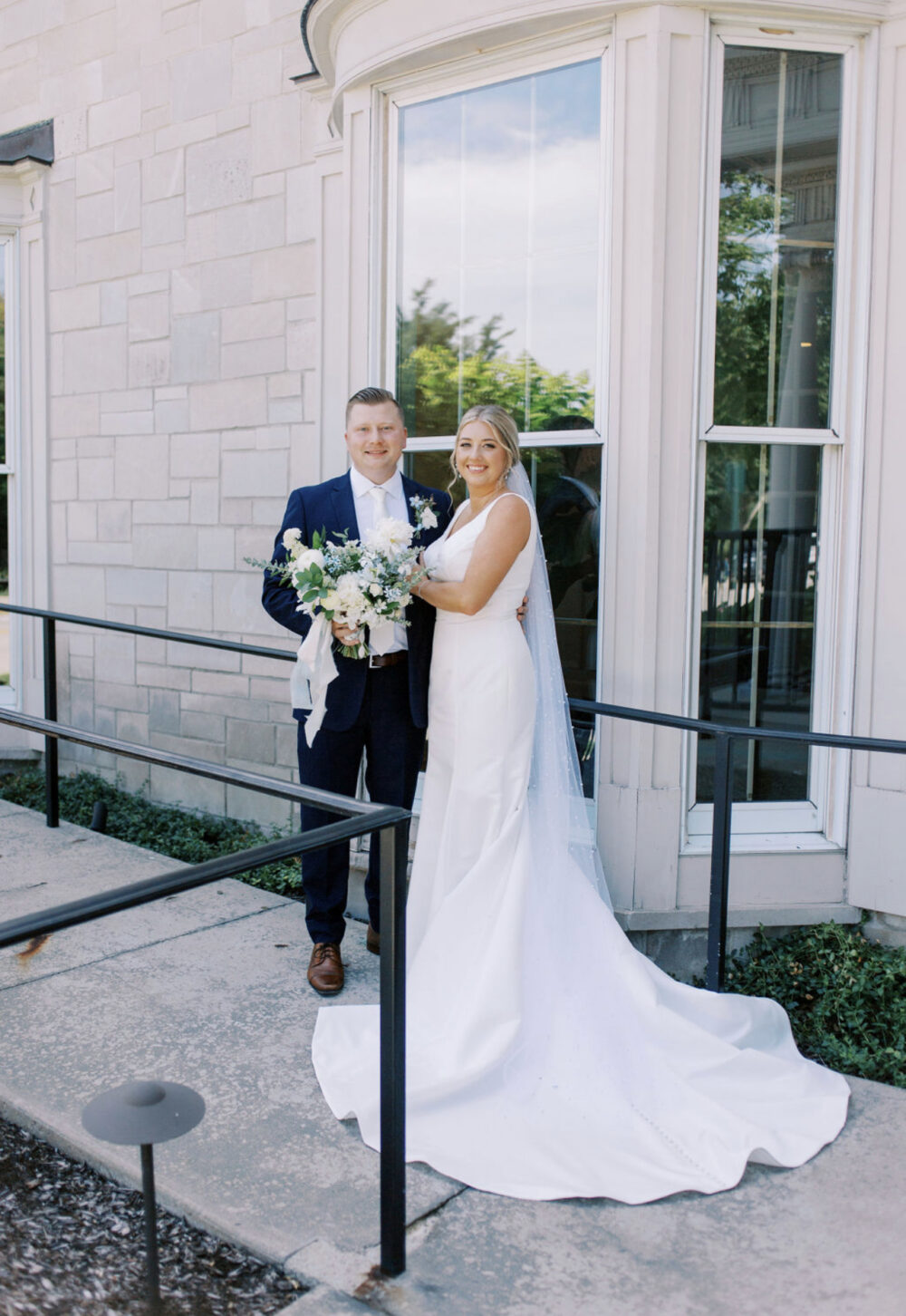 Bride and groom standing together outside a light stone building, holding a bouquet and smiling at the camera.