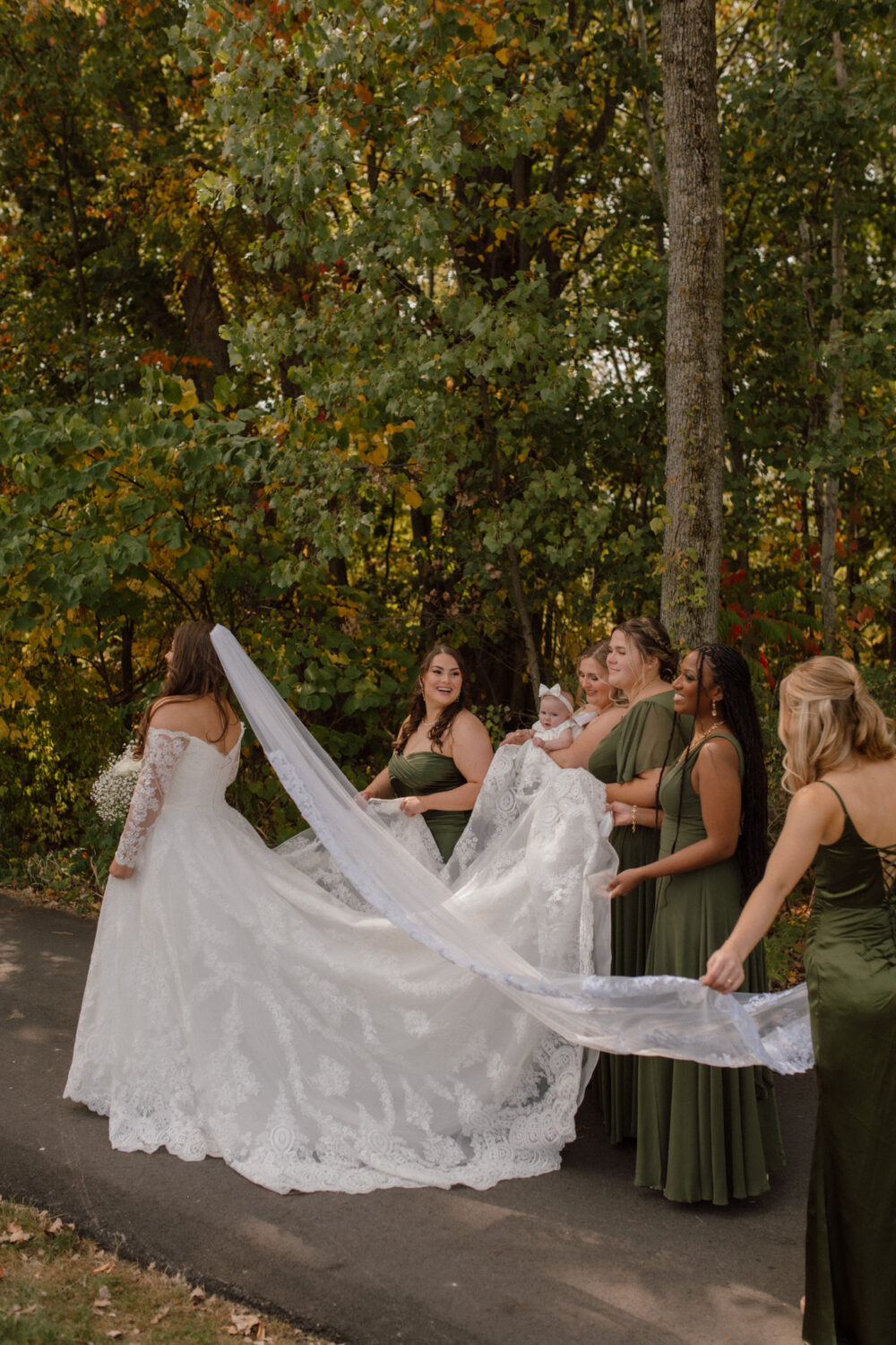 Bride in a lace wedding gown surrounded by bridesmaids in green dresses, holding her train and veil along a wooded pathway.