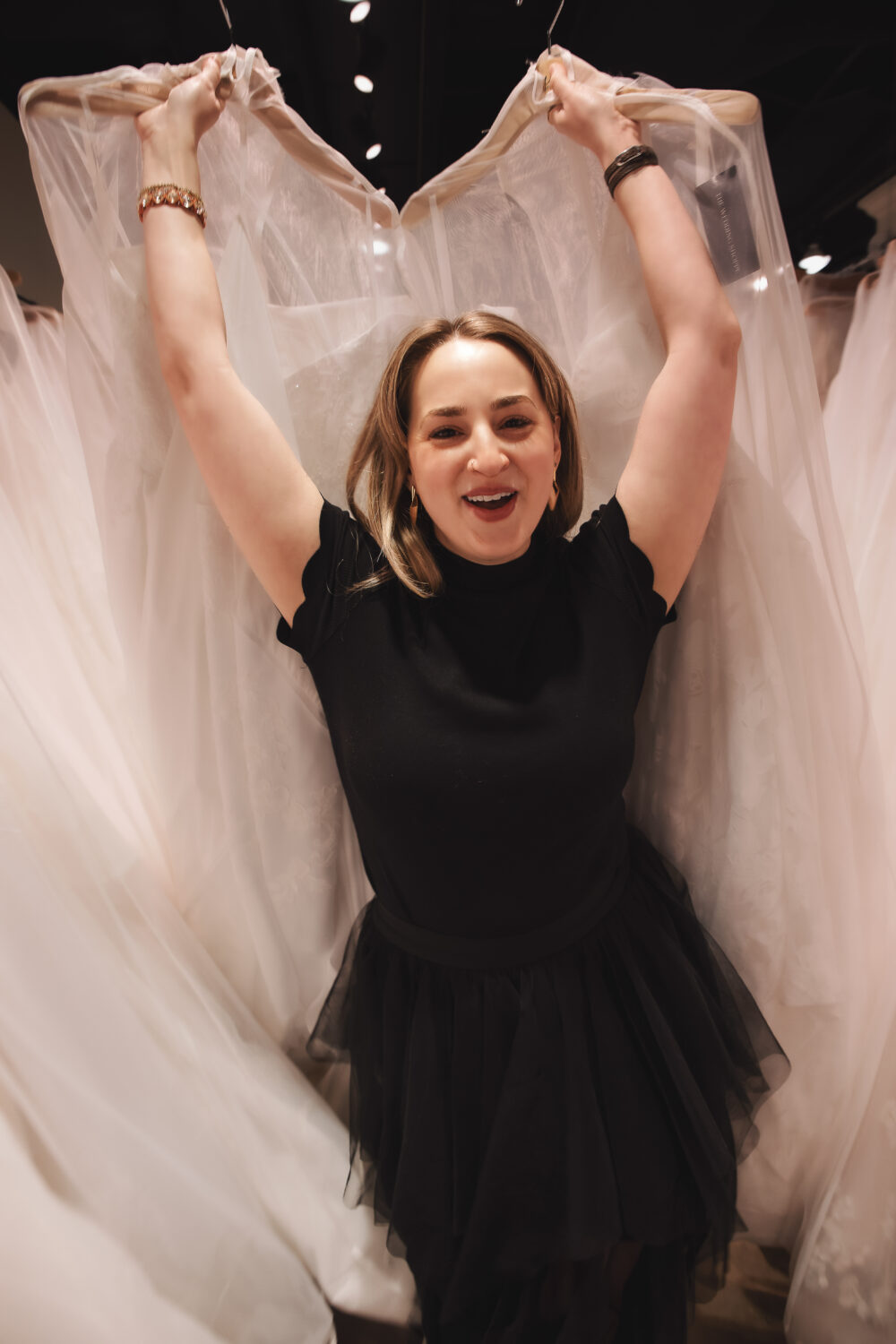 Bridal stylist holding up a wedding gown with a smile while standing between rows of dresses.
