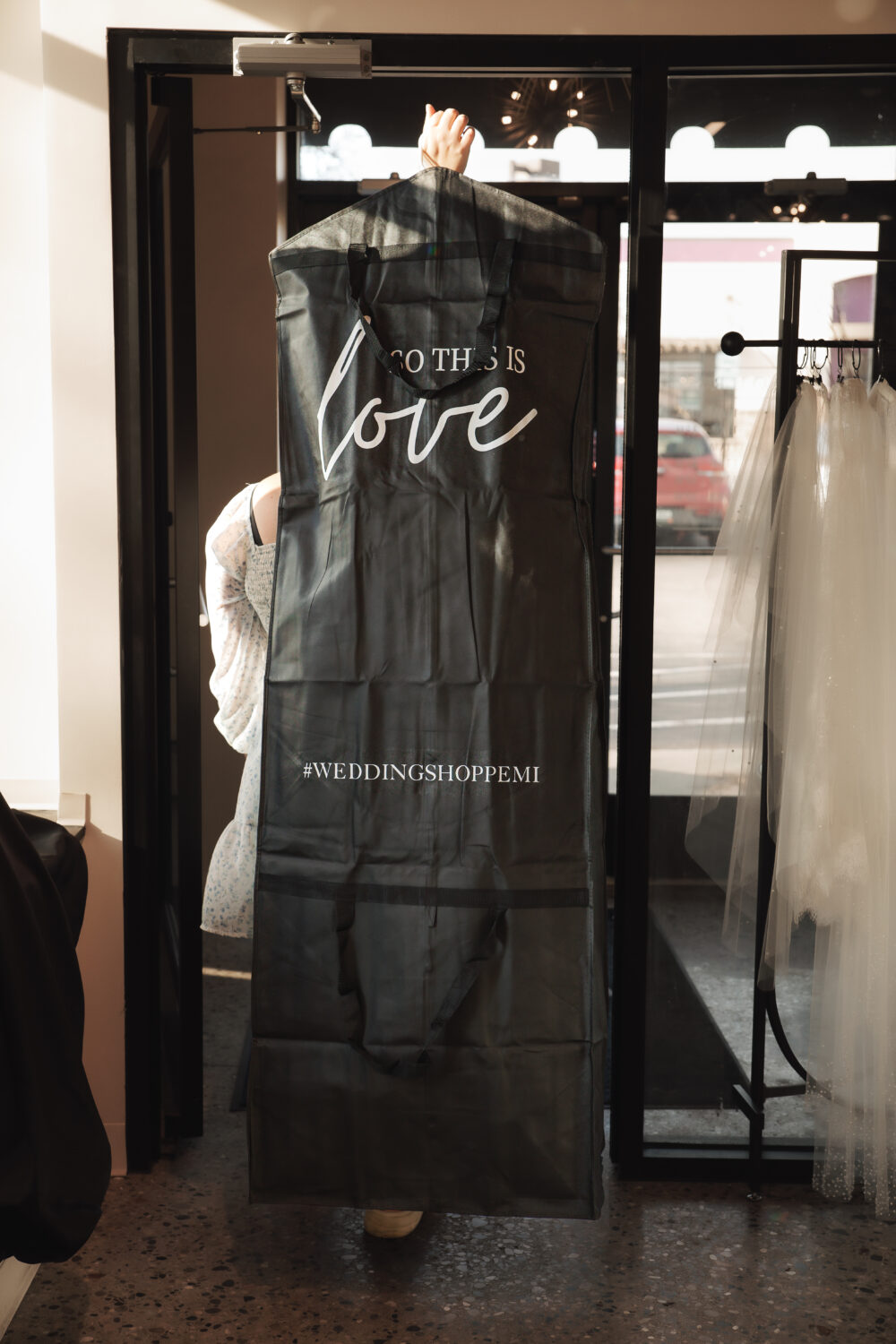 Person carrying a large black garment bag through a boutique entrance with wedding dresses displayed nearby.