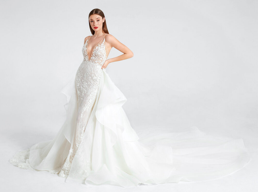 Model in a fitted lace wedding gown with a plunging neckline and flowing overskirt poses against a white studio background.