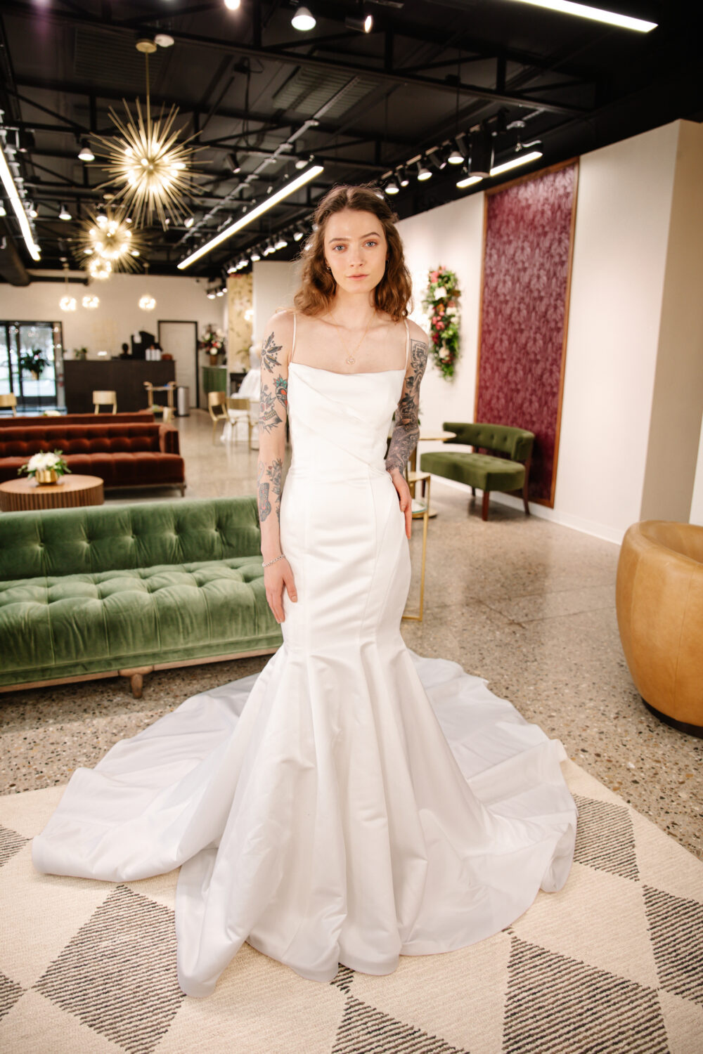 Woman with tattooed arms models a fitted satin mermaid wedding gown with a square neckline and long train inside a bridal boutique.