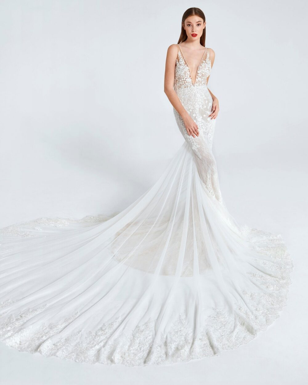 Model wearing a lace mermaid wedding gown with a deep plunging neckline and long tulle train against a white studio backdrop.