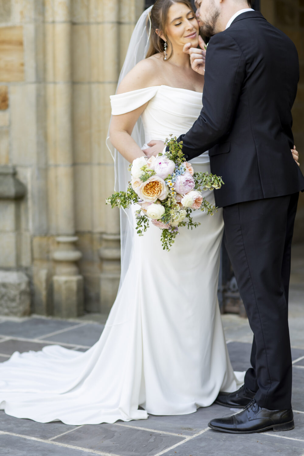 Bride in an off-the-shoulder wedding gown embraces the groom while holding a pastel bouquet outside a stone building.