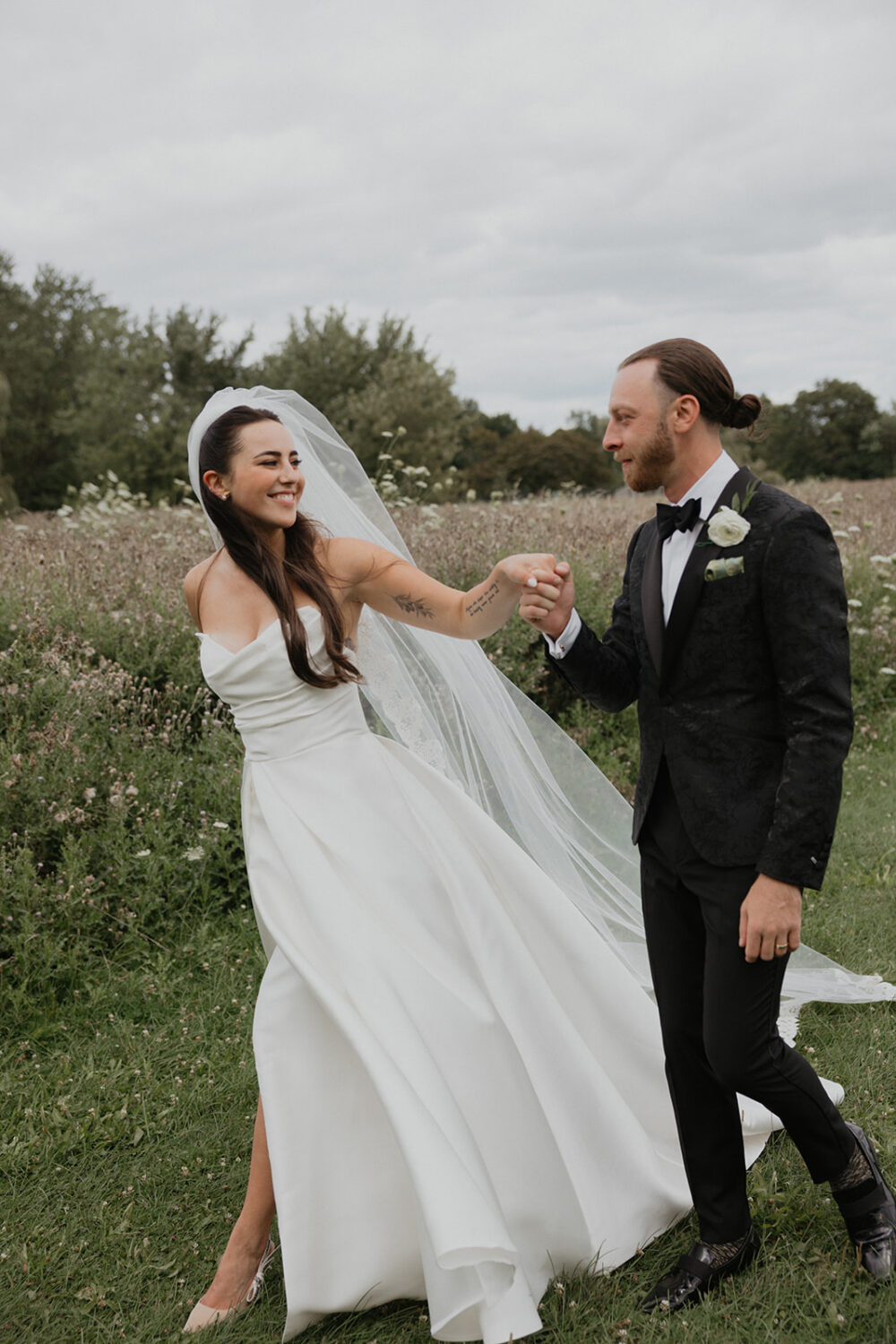 Bride and groom holding hands and smiling at each other while walking through a grassy field in wedding attire.