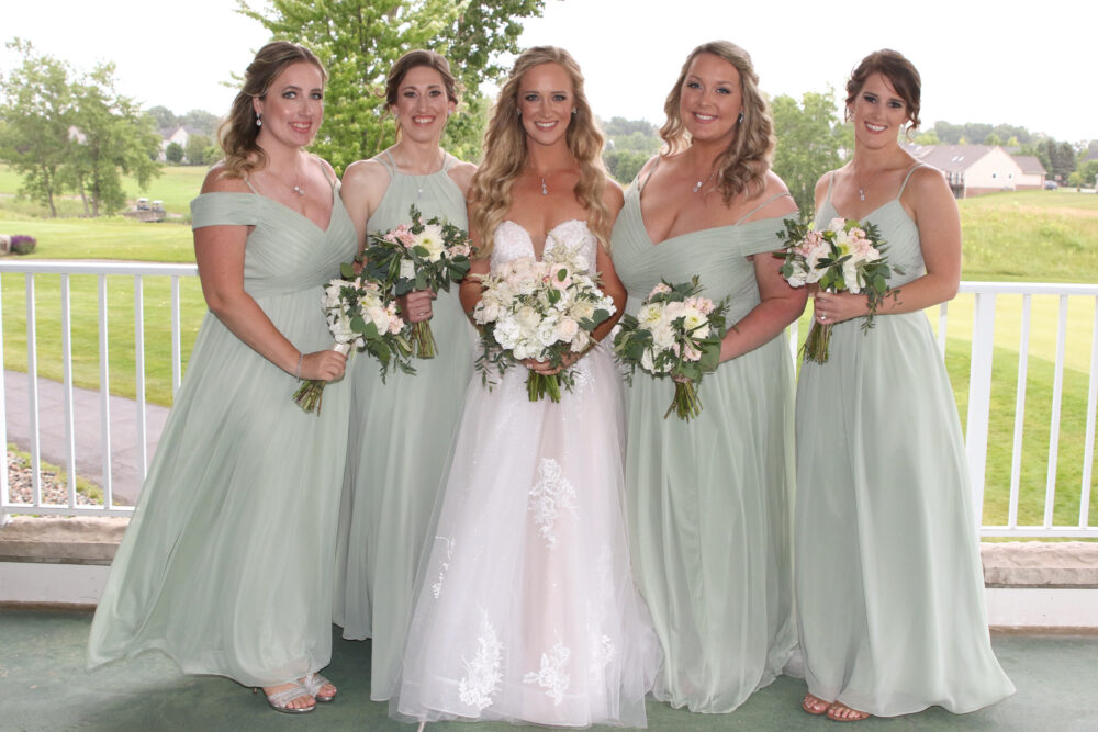 Bride posing with four bridesmaids on a covered porch, each holding bouquets in front of a green landscape.