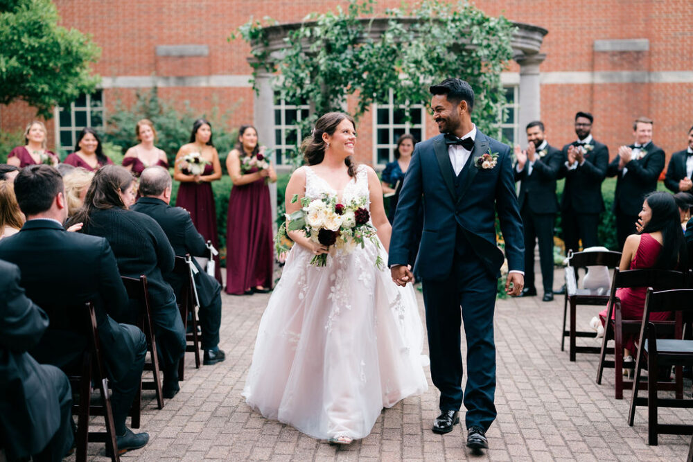 Newly married couple walking down an outdoor aisle together while wedding guests and attendants applaud.
