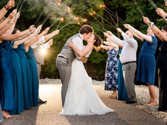 Bride and groom kiss during a sparkler exit as wedding guests line both sides of the walkway holding sparklers.