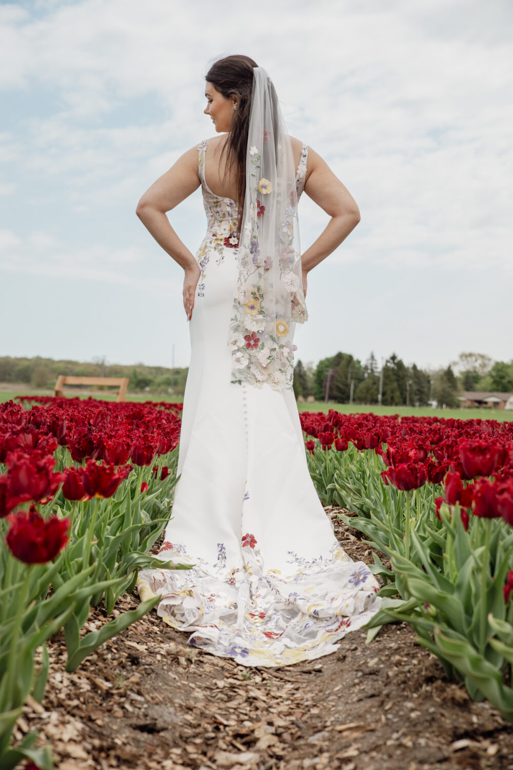 Bride wearing a floral embroidered wedding gown and matching floral veil standing in a field of blooming red tulips.