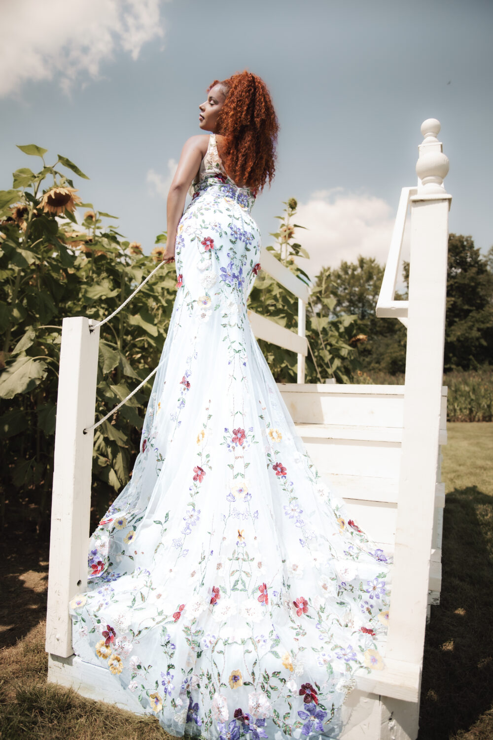 Bride in a floral embroidered wedding dress posing on white wooden steps beside a field of tall sunflowers.
