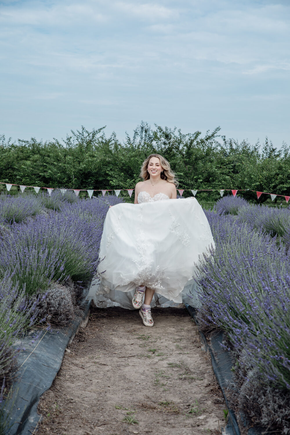 Bride lifting the skirt of her wedding dress while walking through rows of blooming lavender under a cloudy sky.