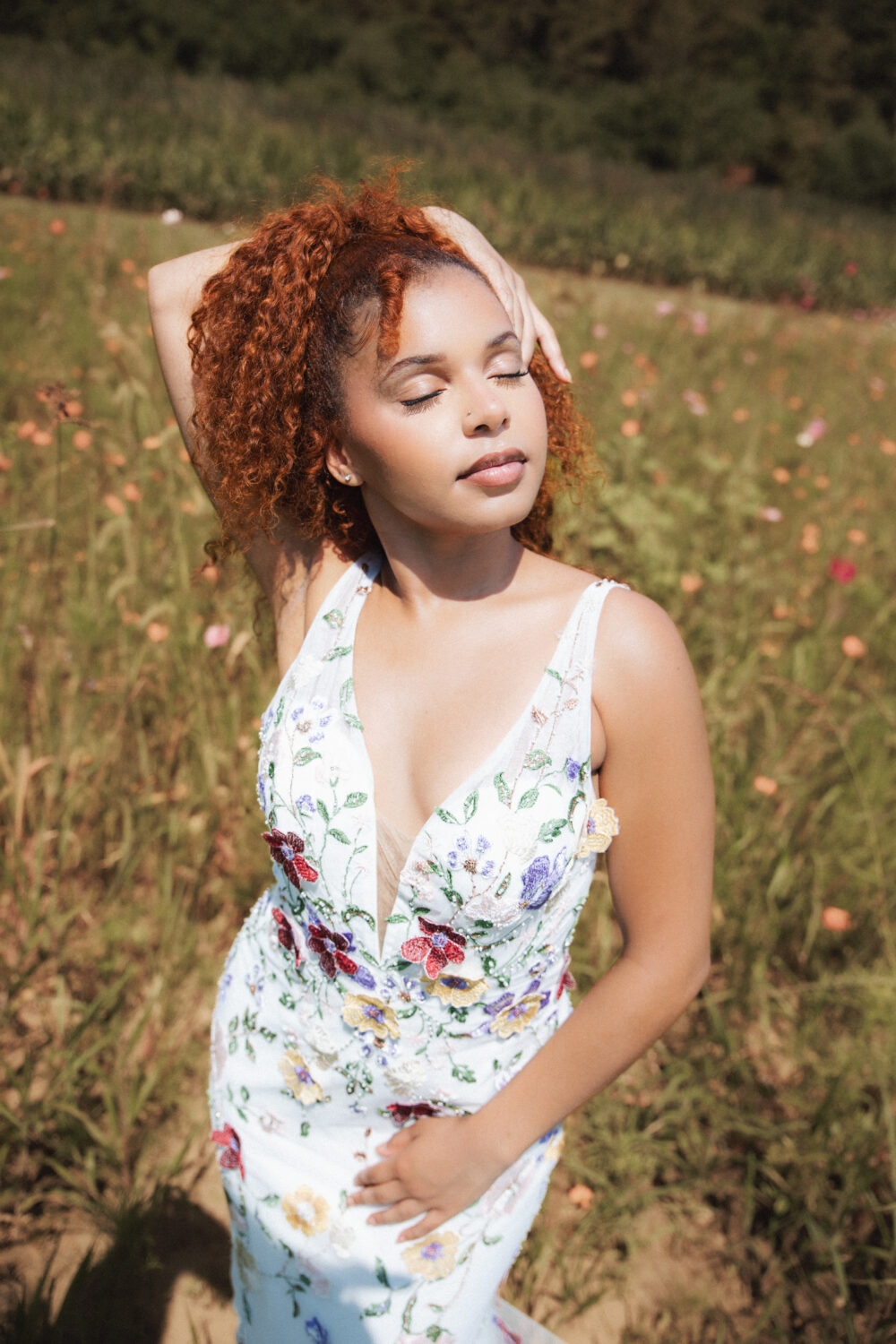 Bride with curly red hair posing in a floral embroidered wedding gown in a sunlit wildflower meadow.