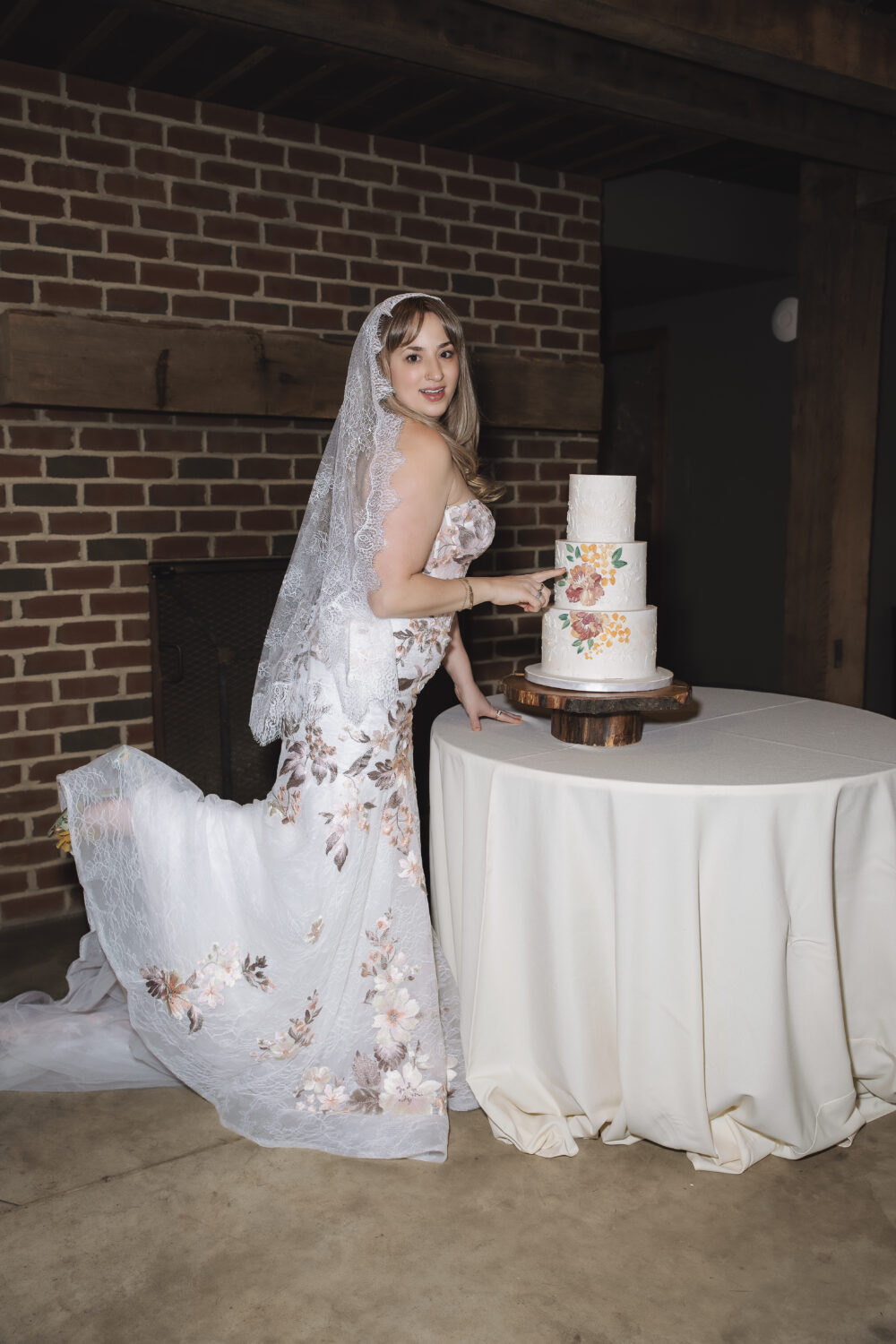 Bride in a floral lace wedding gown and veil posing beside a three-tier floral wedding cake in a rustic indoor venue.