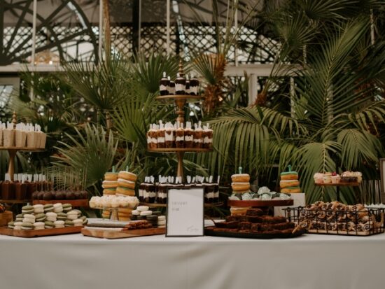 A wedding dessert display features assorted cakes, cookies, macarons, brownies, and pastries arranged on tiered stands in a lush indoor garden setting.