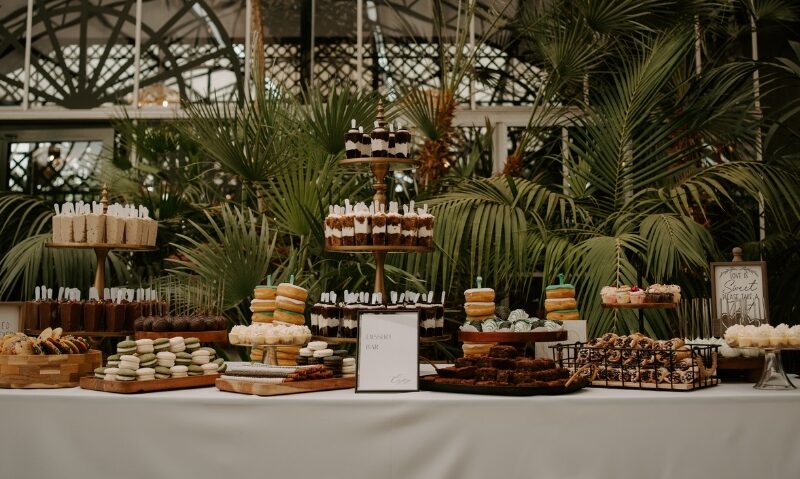 A wedding dessert display features assorted cakes, cookies, macarons, brownies, and pastries arranged on tiered stands in a lush indoor garden setting.
