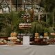 A wedding dessert display features assorted cakes, cookies, macarons, brownies, and pastries arranged on tiered stands in a lush indoor garden setting.