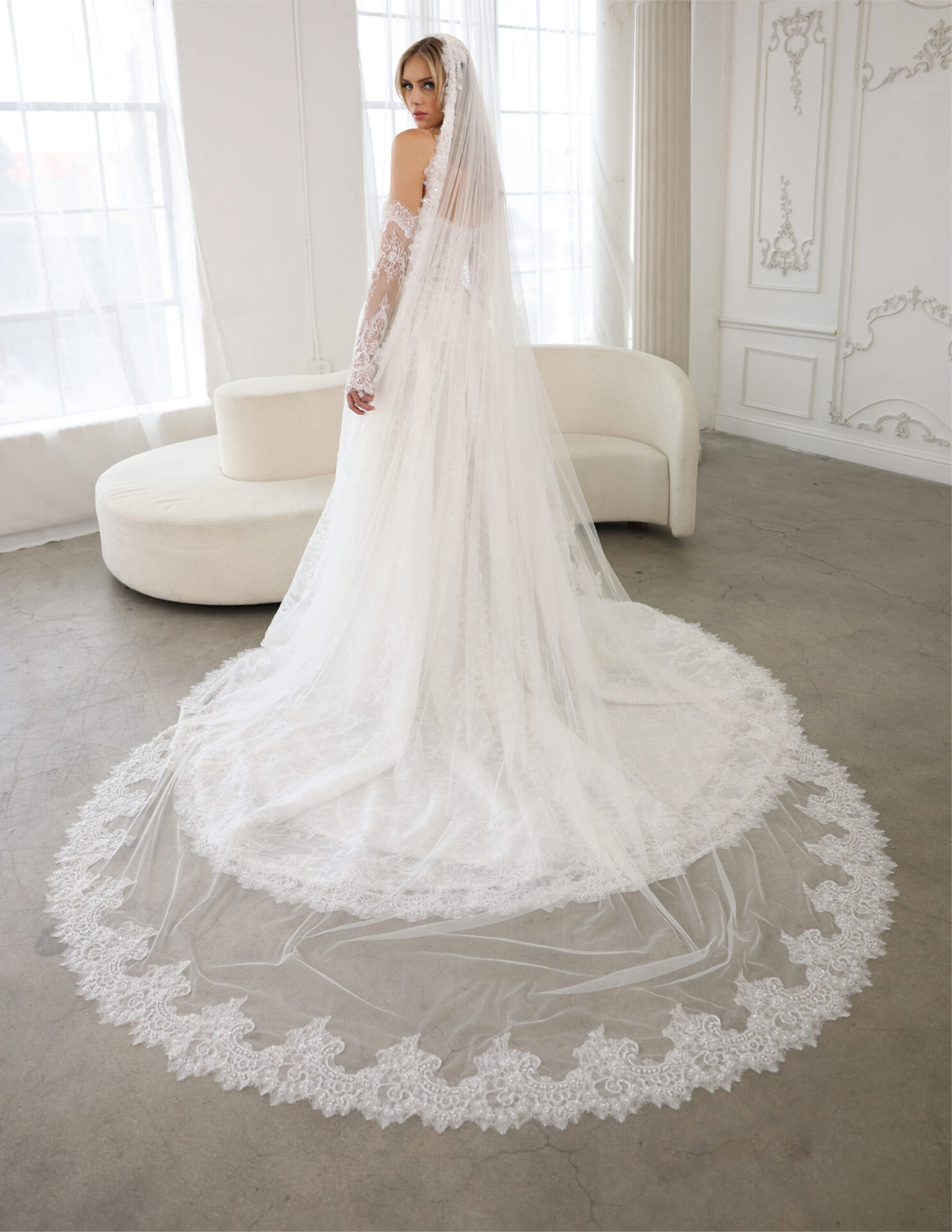 Bride wearing a lace wedding gown with a dramatic cathedral-length veil and long lace train in a bright bridal studio.