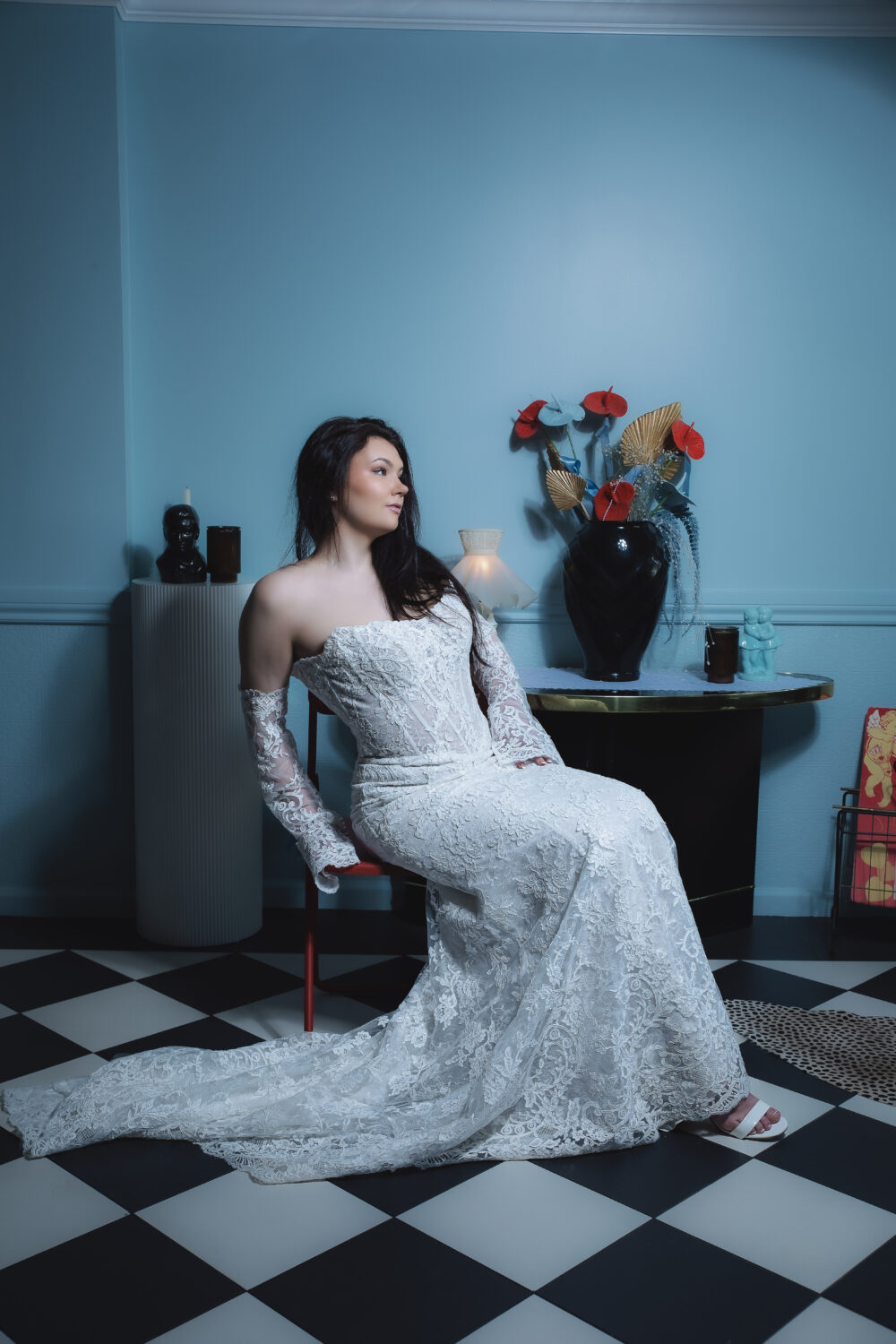 A bride in an off-the-shoulder lace wedding gown with sheer lace sleeves sits on a red chair in a stylish room with black-and-white checkered flooring, decorative vases, and a long lace train.
