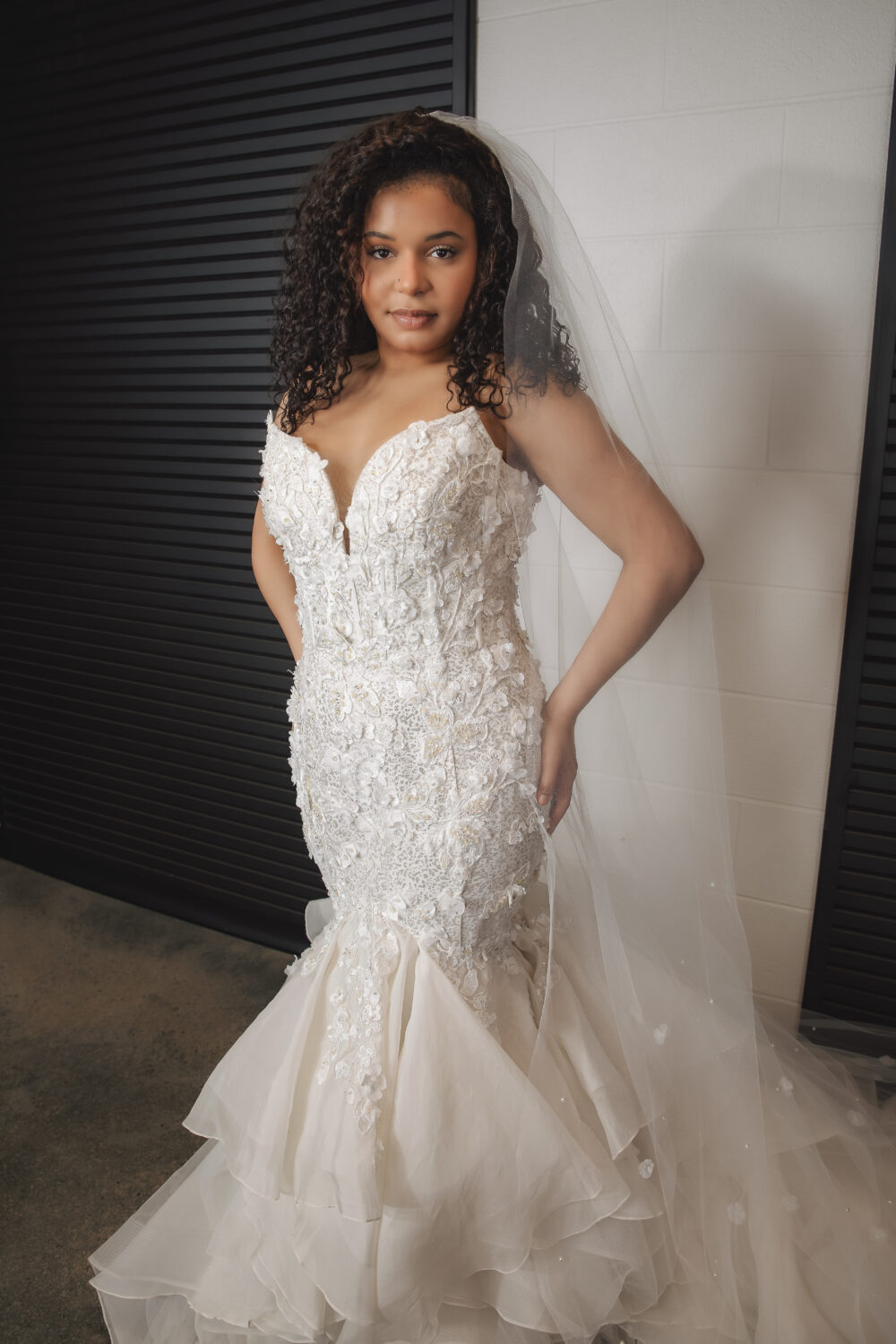 A bride wearing a fitted floral lace mermaid wedding gown with a sweetheart neckline and a long veil poses indoors in front of black shutter doors.