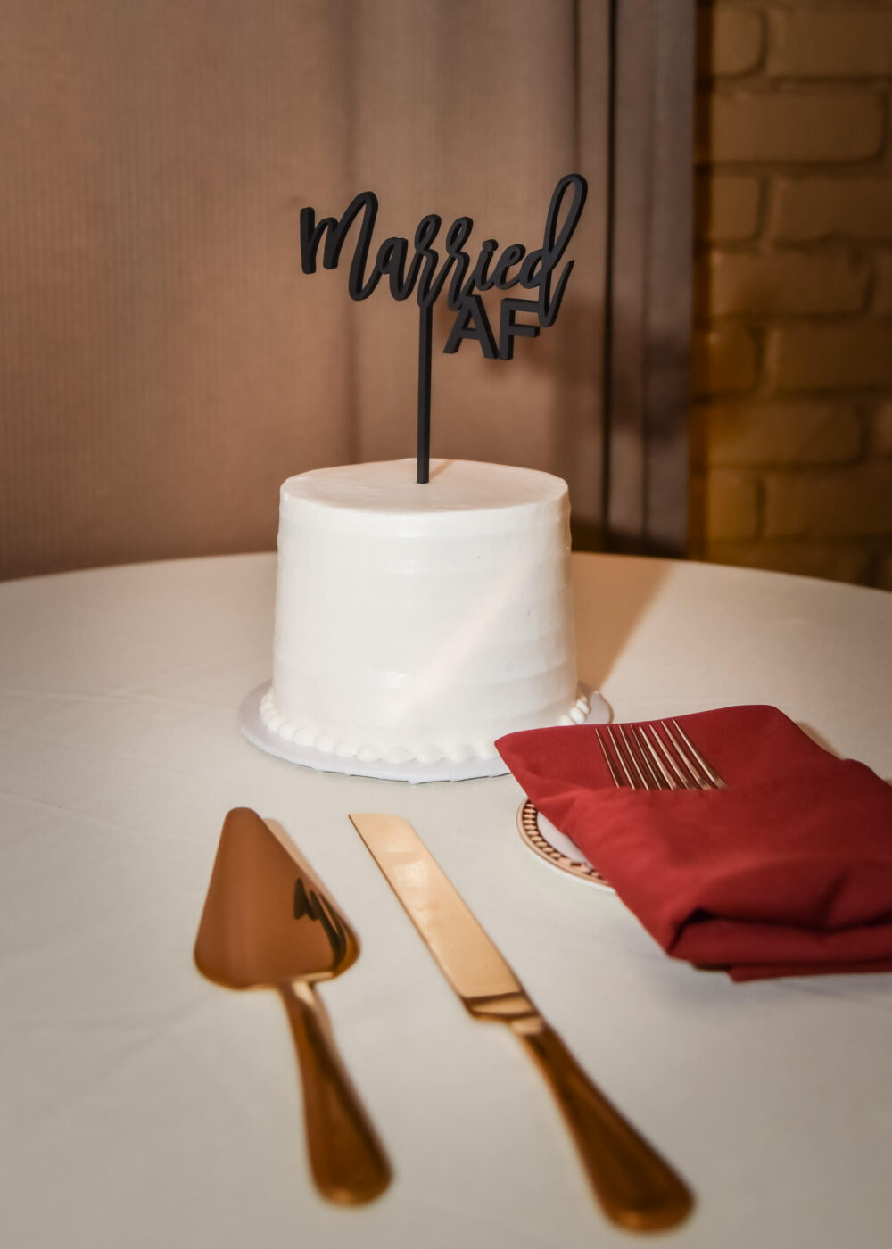 A small white wedding cake with a black "Married AF" topper sits on a table beside a cake server, knife, and folded burgundy napkin.