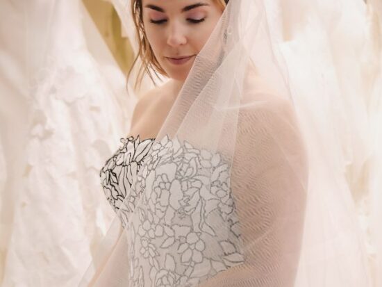 A bride wearing a strapless floral-pattern wedding gown and a sheer veil looks down while standing among rows of hanging wedding dresses.