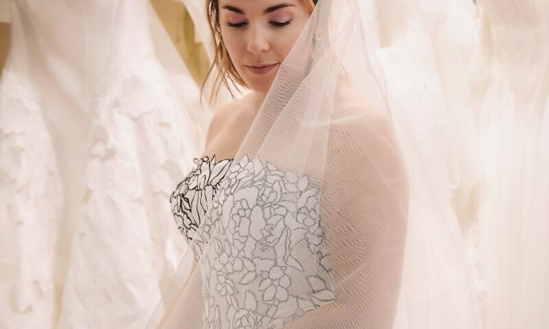 A bride wearing a strapless floral-pattern wedding gown and a sheer veil looks down while standing among rows of hanging wedding dresses.