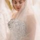 A bride wearing a strapless floral-pattern wedding gown and a sheer veil looks down while standing among rows of hanging wedding dresses.