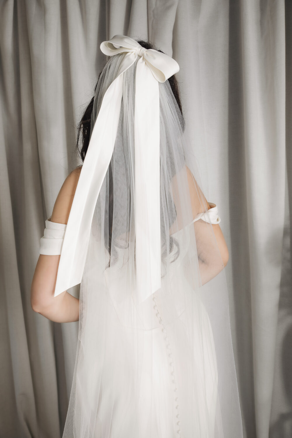 The back of a bride wearing an off-the-shoulder wedding gown with covered buttons, a long veil, and a large satin bow in her hair stands in front of light-colored curtains.