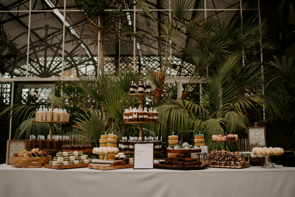 A wedding dessert display features assorted cakes, cookies, macarons, brownies, and pastries arranged on tiered stands in a lush indoor garden setting.