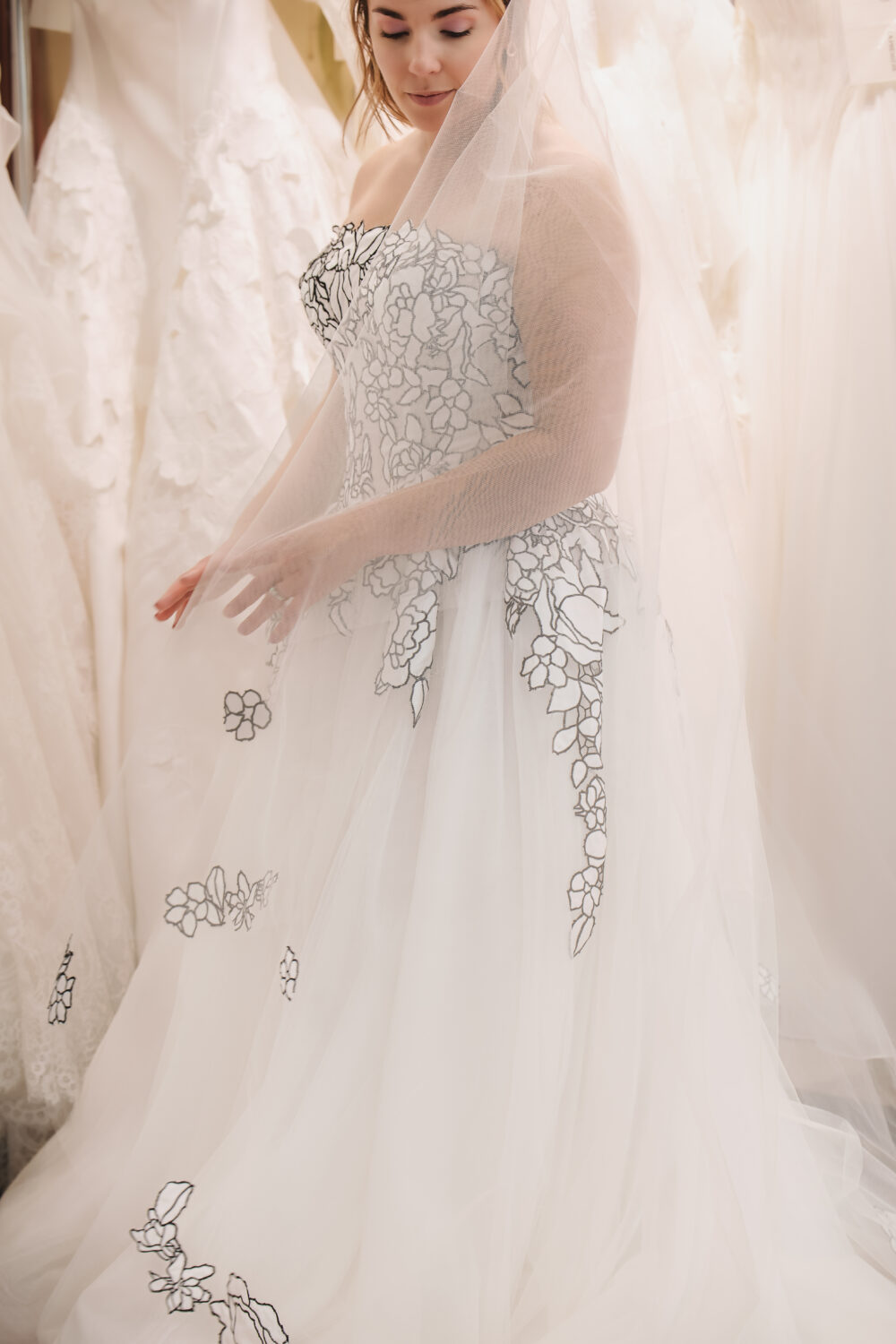 A bride wearing a strapless floral-pattern wedding gown and a sheer embroidered veil gently holds the veil while standing among hanging wedding dresses.