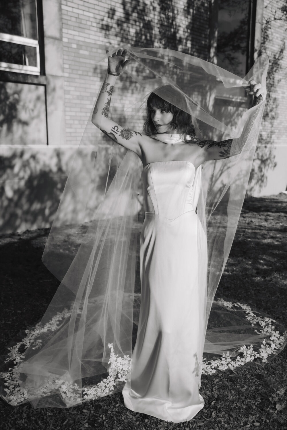 A bride with tattooed arms wears a minimalist strapless wedding gown and lifts a long sheer veil above her head while standing outdoors in black and white.