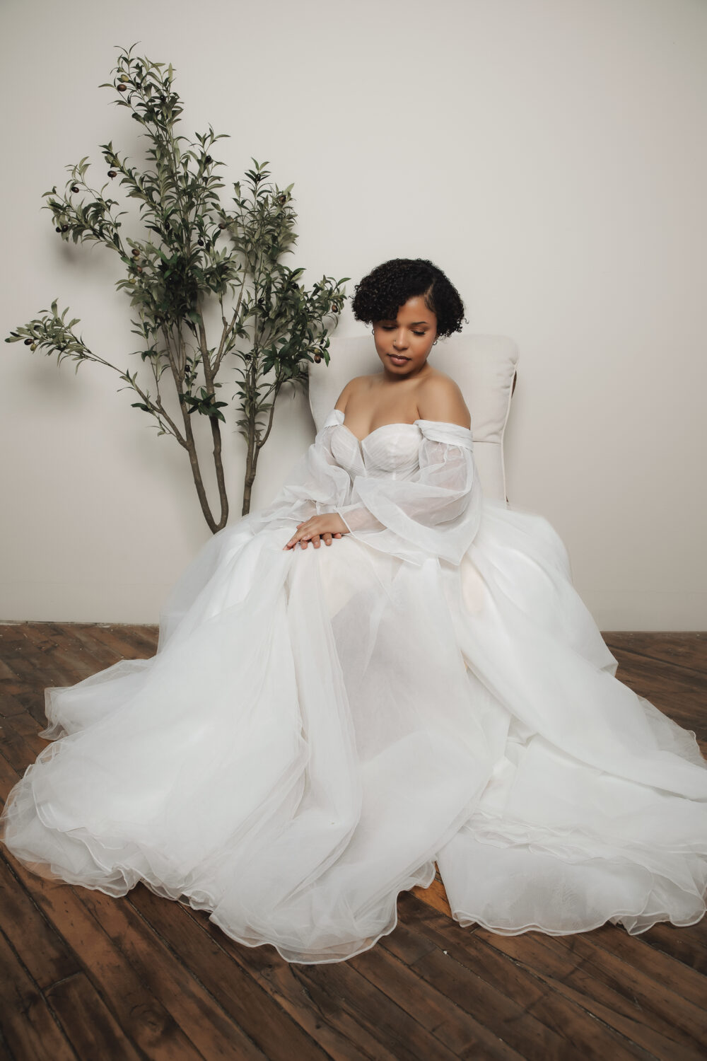 A bride wearing an off-the-shoulder tulle ball gown sits in a white chair with her hands folded in her lap beside a decorative indoor tree.