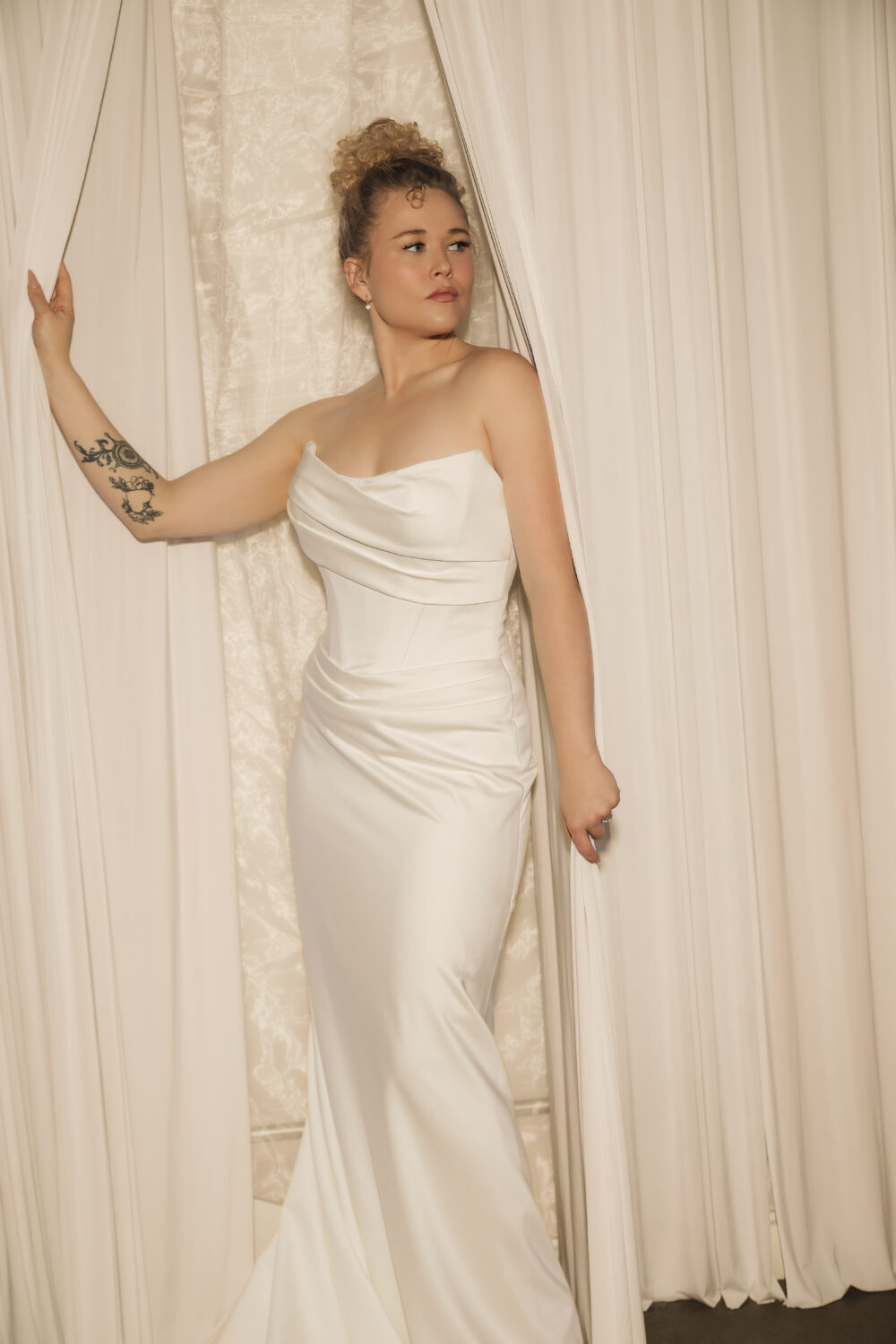 A bride wearing a sleek strapless satin wedding gown stands between flowing white curtains, looking to the side with one hand resting on the drapery.