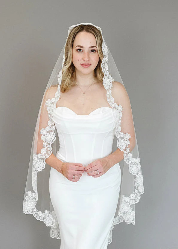 Bride in a fitted satin wedding dress wearing a sheer veil with floral lace trim against a plain gray background.