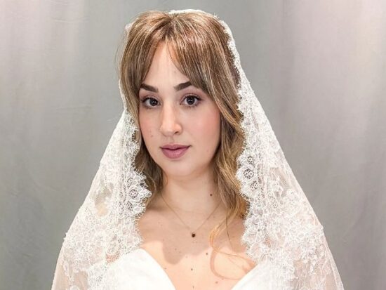 Close-up portrait of a bride wearing a lace-edged veil over a satin wedding dress against a gray backdrop.