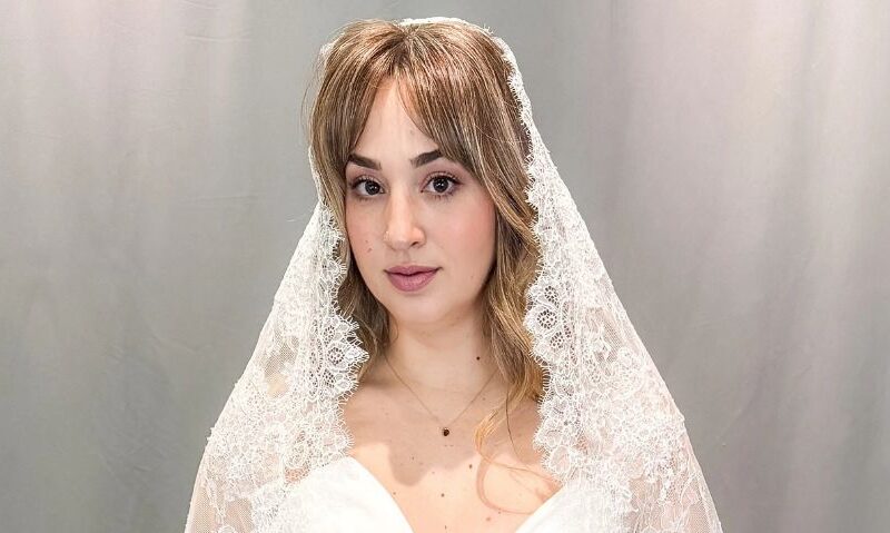 Close-up portrait of a bride wearing a lace-edged veil over a satin wedding dress against a gray backdrop.
