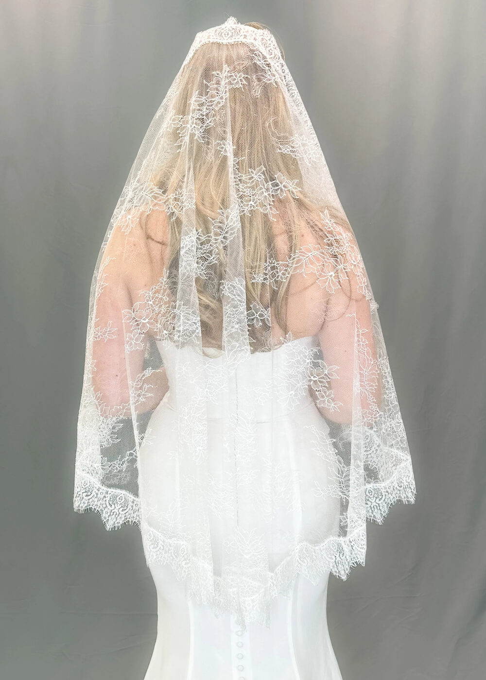 Back view of a bride wearing a long sheer veil with delicate floral lace edging over a fitted white wedding dress.