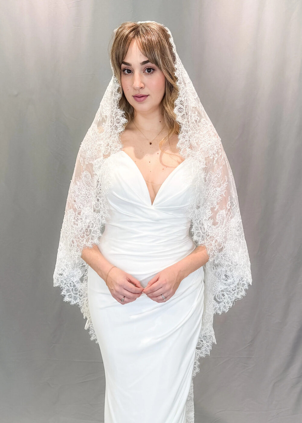 Bride wearing a satin wedding dress with a lace-edged veil draped over her head and shoulders against a gray backdrop.
