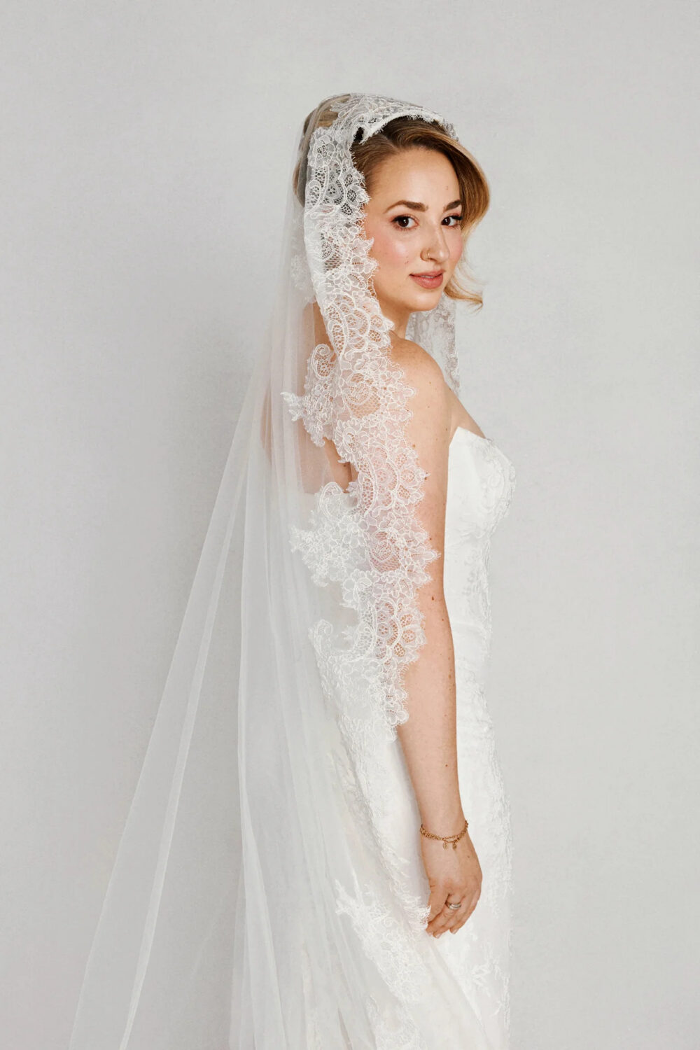 Side profile of a bride in a strapless lace wedding dress wearing a long lace-edged veil and looking over her shoulder.