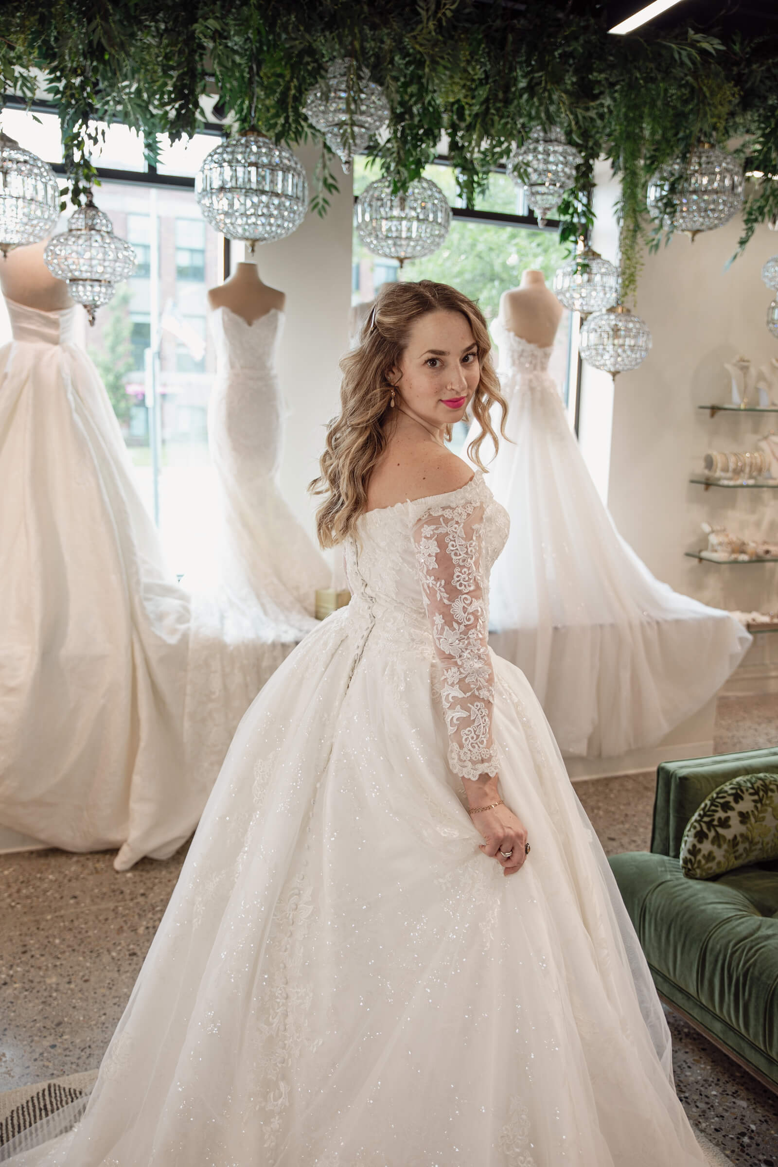 Bride in a long-sleeve lace ball gown posing inside a bridal boutique surrounded by wedding dresses.