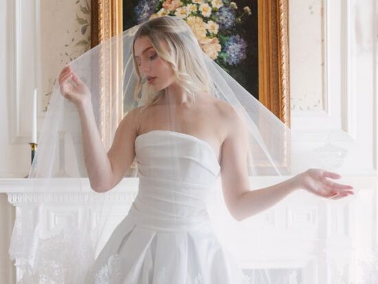 Bride in a strapless satin wedding gown holds the edges of her veil while posing in front of a fireplace with a floral painting above it.