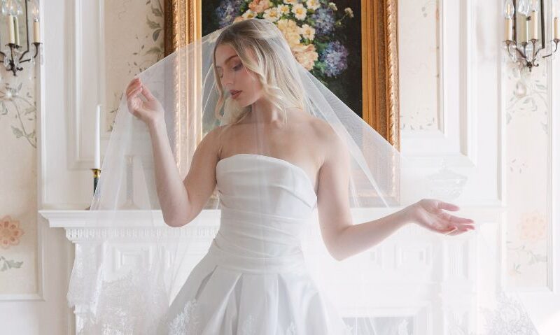 Bride in a strapless satin wedding gown holds the edges of her veil while posing in front of a fireplace with a floral painting above it.
