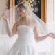 Bride in a strapless satin wedding gown holds the edges of her veil while posing in front of a fireplace with a floral painting above it.