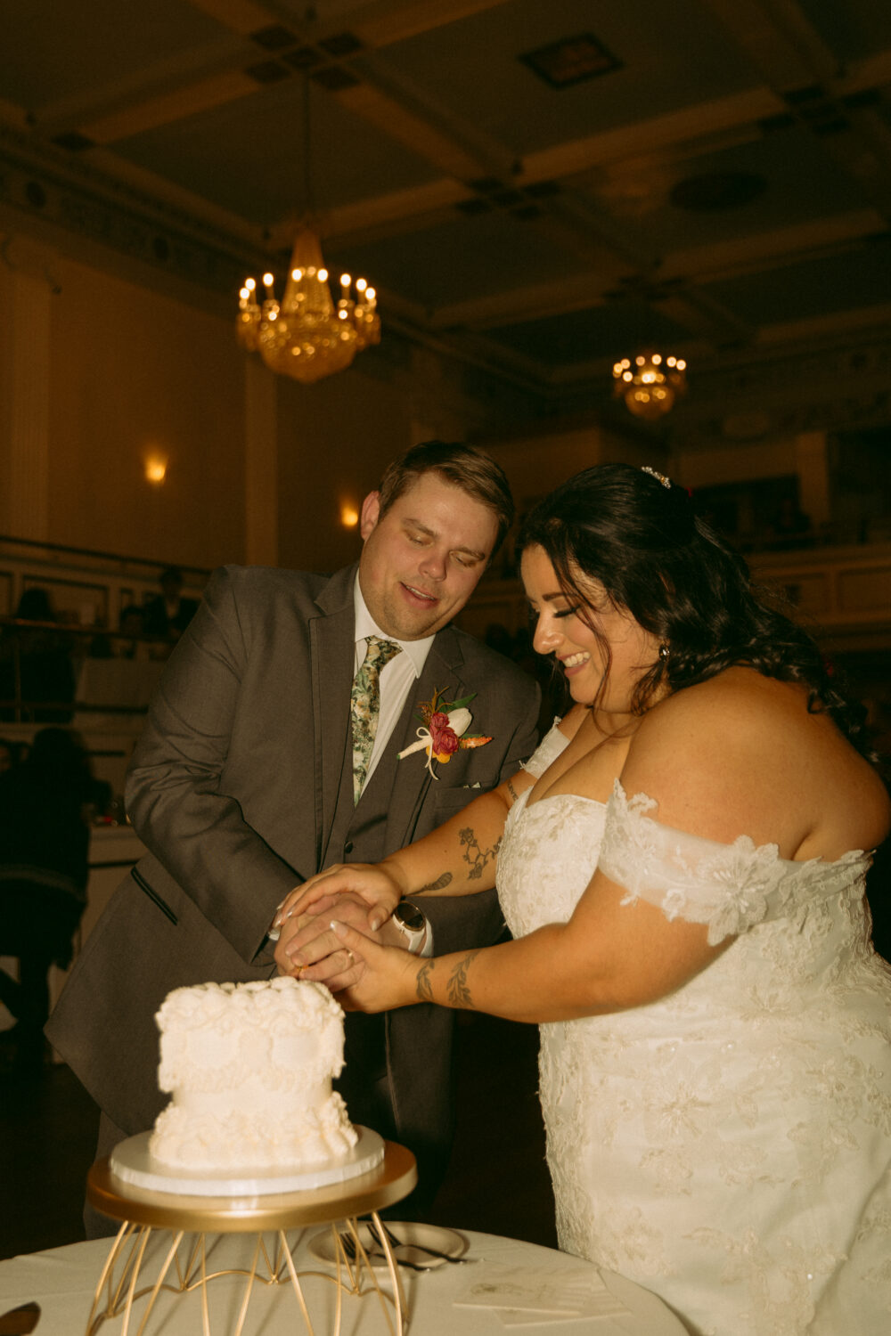 A bride and groom cut a white wedding cake together at their reception while smiling and standing beside a gold cake stand in a warmly lit ballroom.