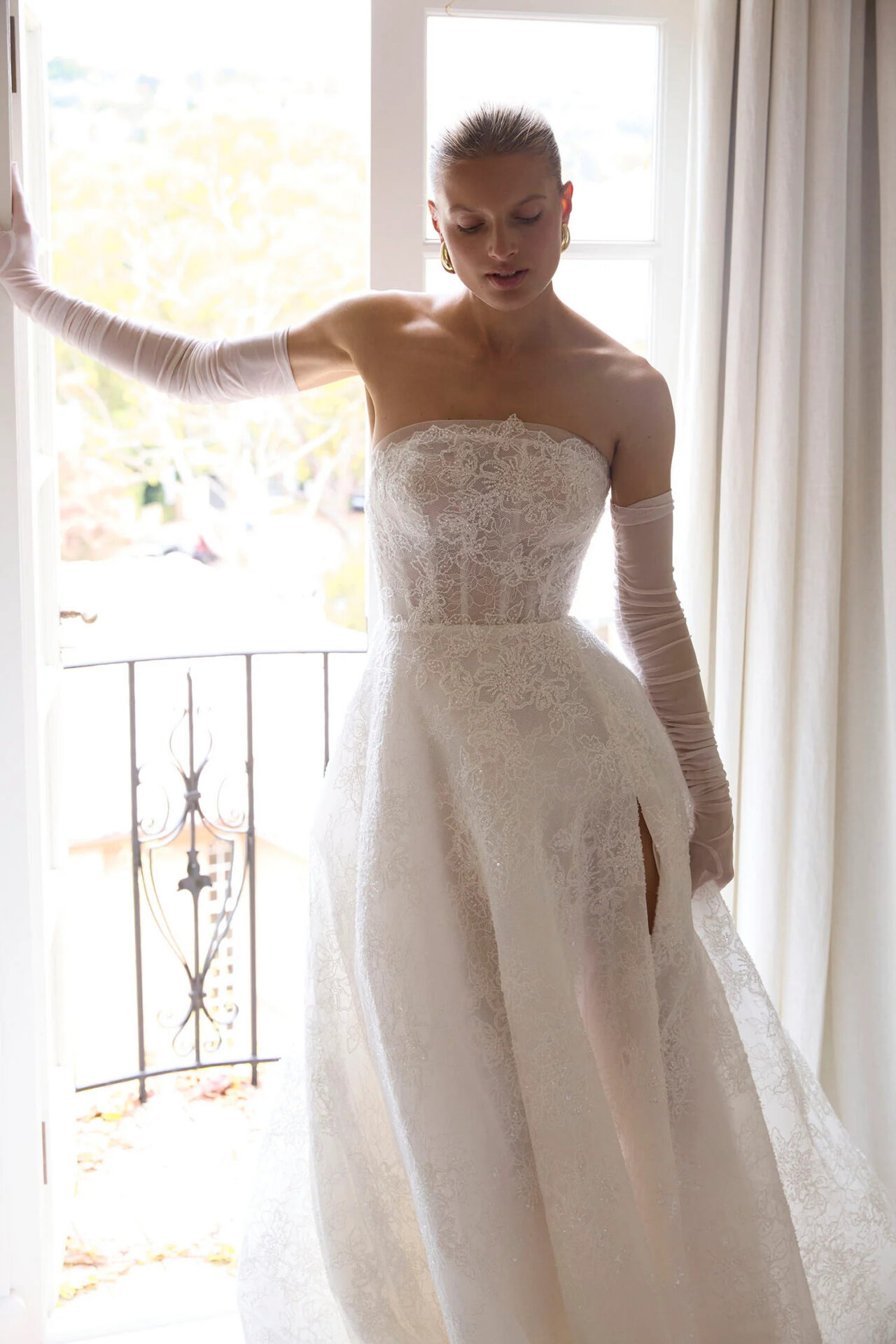 Bride wearing a strapless lace wedding gown with matching sheer opera gloves beside a sunlit window.